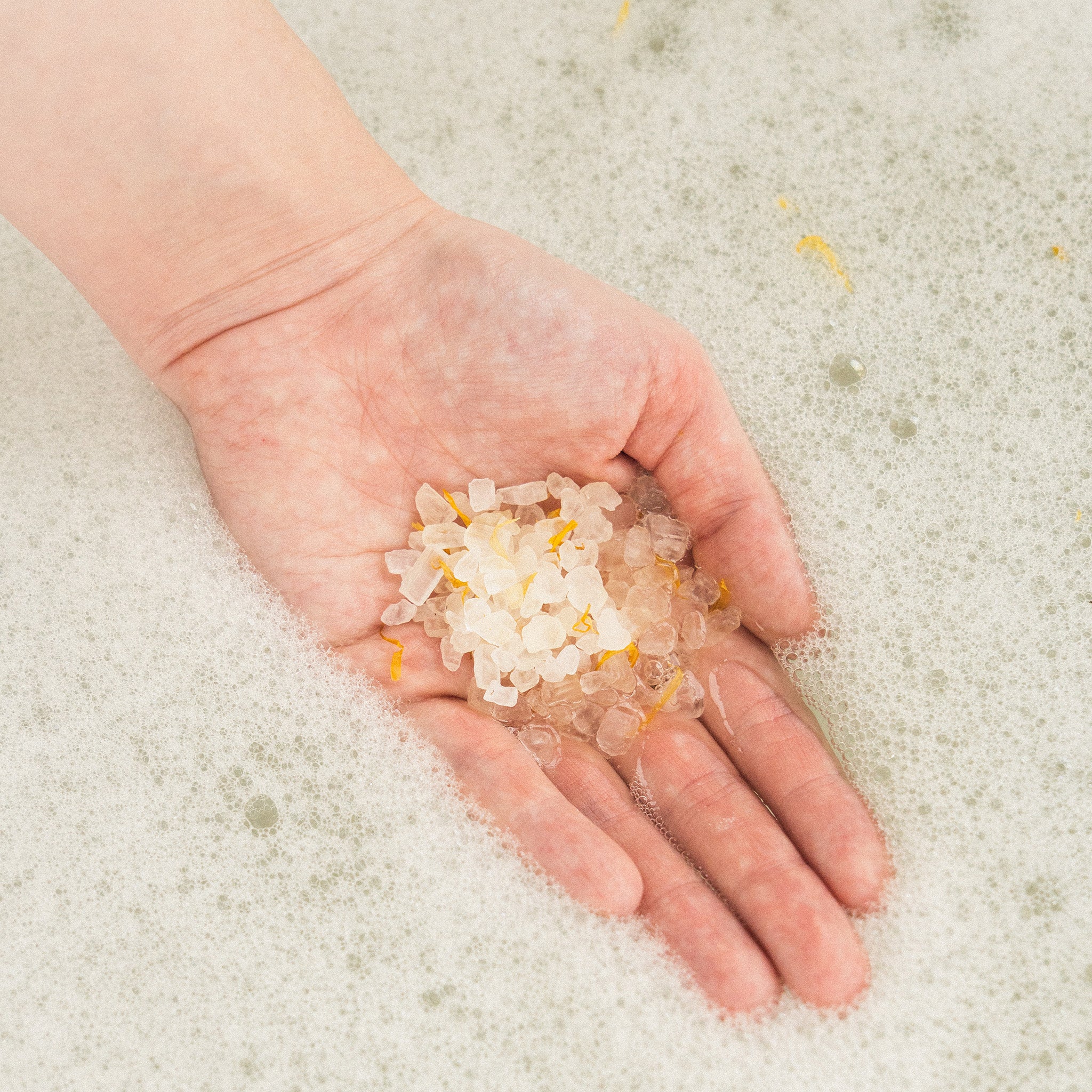 Dłoń trzymająca przezroczyste kryształki soli do kąpieli z dodatkiem złotych płatków, na tle białej, pienistej wody.