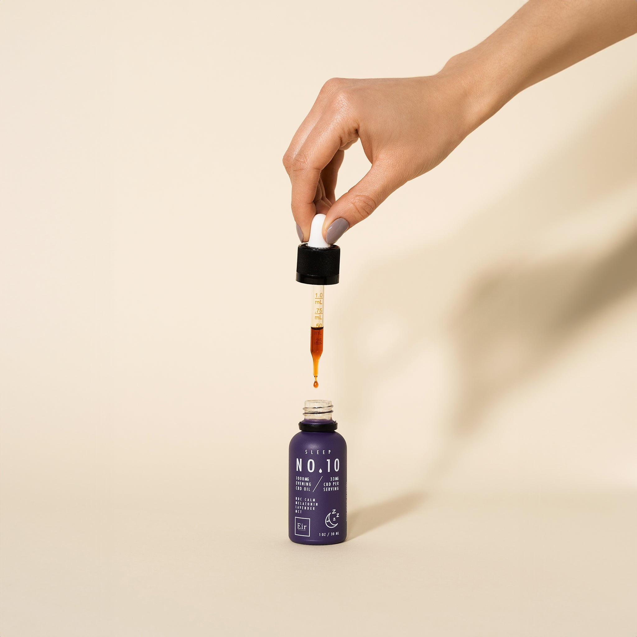 Dłoń trzymająca zakraplacz nad fioletową butelką olejku CBD Eir No. 10, z kroplą oleju zawieszoną na końcu, na neutralnym tle.