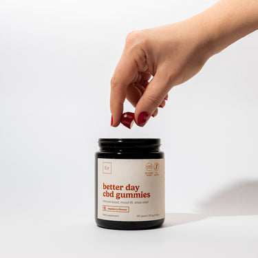 Dłoń z czerwonym manicure sięgająca po słoik żelków CBD Better Day. Czarne opakowanie z białą etykietą na jasnym tle, pokazujące codzienny sposób użycia suplementu.